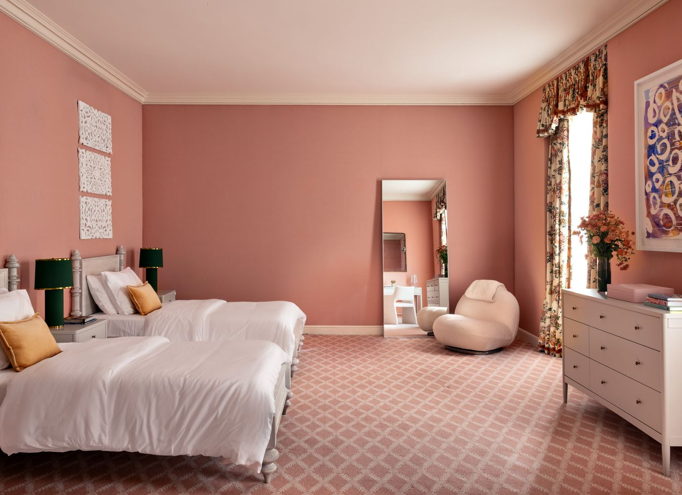 Bedroom painted in dusty pink walls and patterned carpeting, with two twin beds facing the floral draped windows.