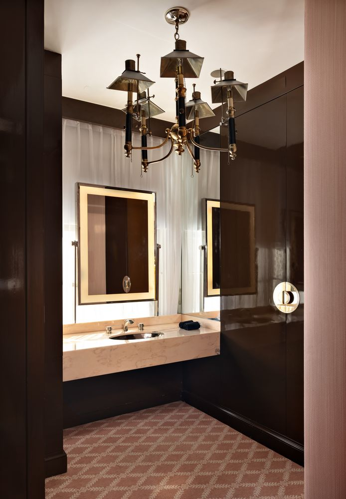 Its en-suite bathroom has glossy brown wall closets surrounding a floating stone vanity and a large illuminated mirror beneath a brass chandelier.