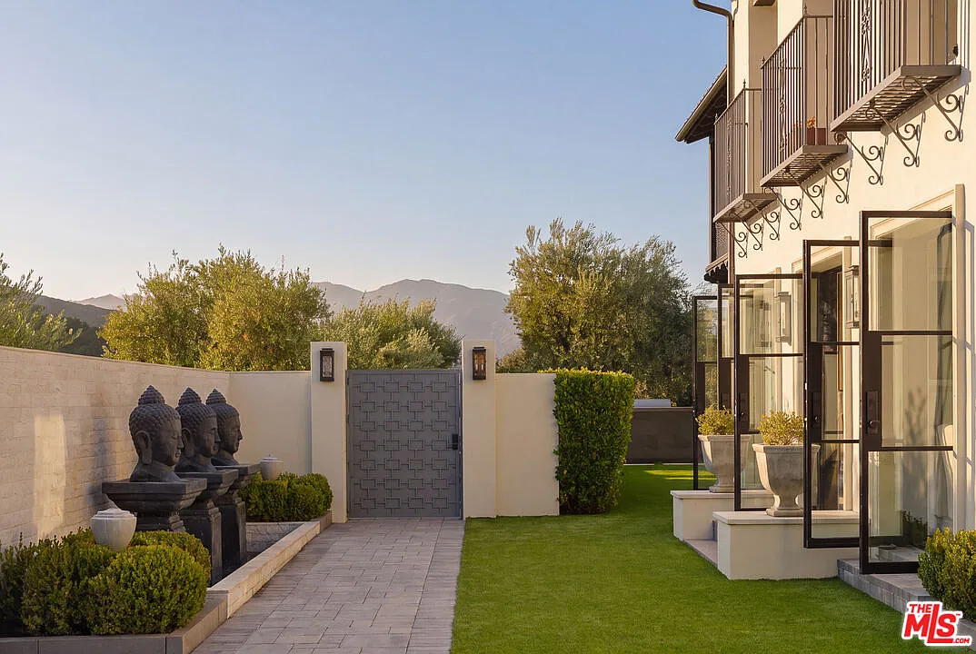A private gate opens to a landscaped walkway that runs alongside the house toward the front door, with clipped greenery and statues lining the path.